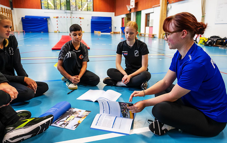 Bilden visar ungdomar som sitter i en ring i en gympasal där ledaren bläddrar in en broschyr